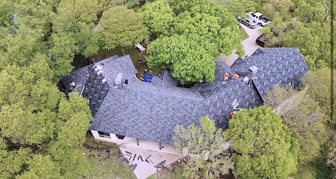 Drone view of a roofing crew laying charcoal shingles on a house surrounded by trees